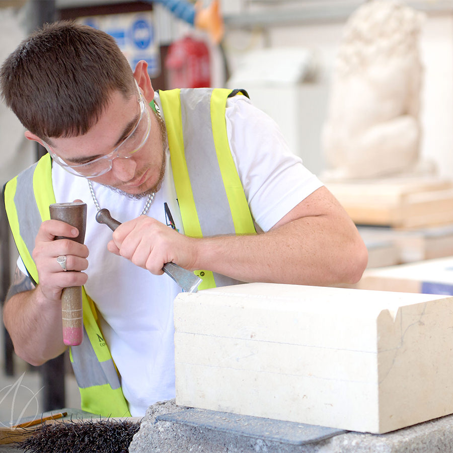 Apprentice stonemason carving a limestone block with chisel and mallet, wearing safety glasses and hi-vis vest
